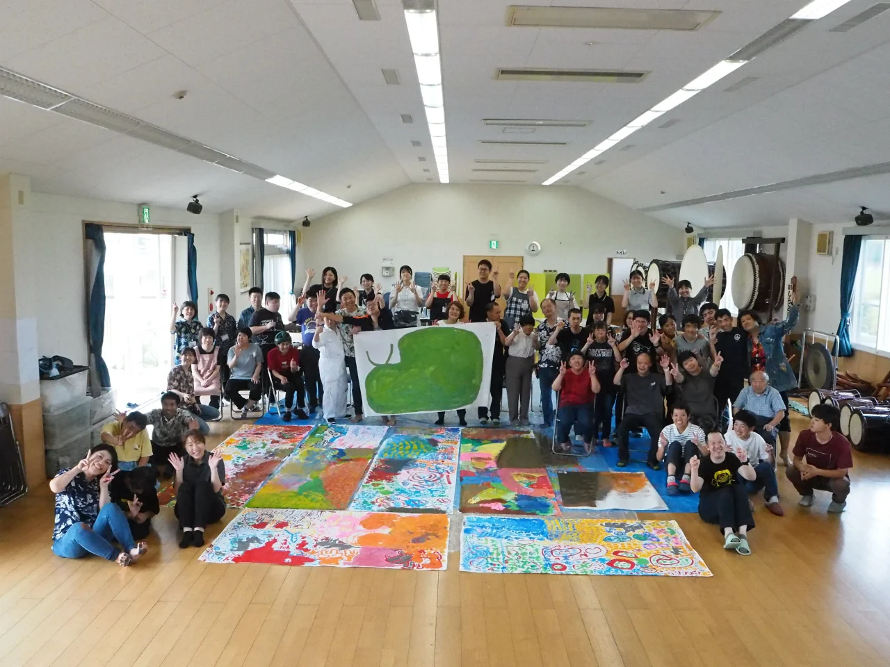 徳武産業・德武社長（中央）と、アートを描き上げた征峯会の利用者様、スタッフ一同。大きな紙を囲んで共に色を重ねたあの日、私たちは「支援する側・される側」という垣根を超え、一つのチームになりました。