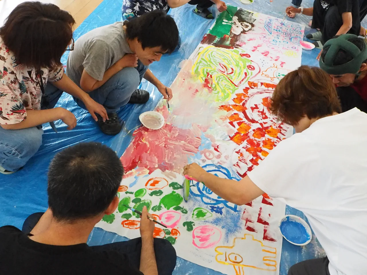 大きな白布を囲み、德武社長（右側手前）と利用者様が共に筆を走らせる様子。そこには「企業と福祉」という垣根はなく、ただ純粋に表現を楽しむ、一人の人間同士の豊かな時間が流れていました。