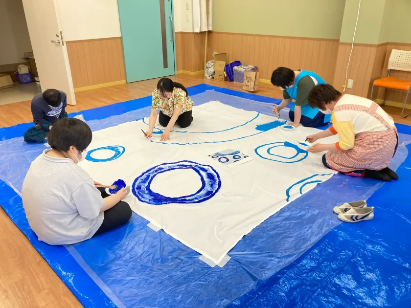 ブルーシートの上で、数人の利用者が大きな布に青い絵の具で円や模様を描いている様子
