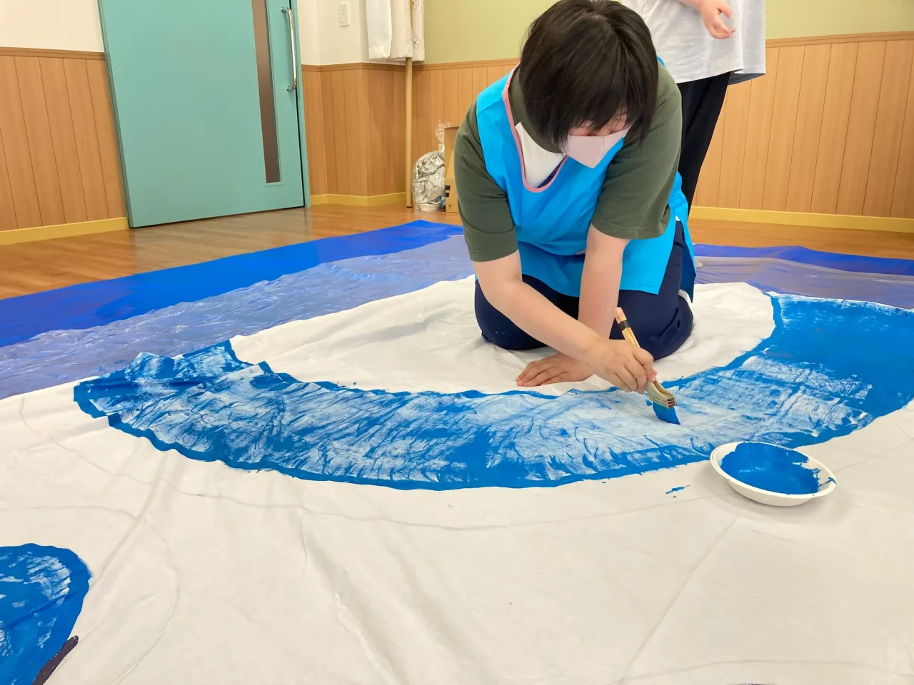 大きな白い布にしゃがみ込み、太い筆を使って青い絵の具を丁寧に塗る利用者の様子