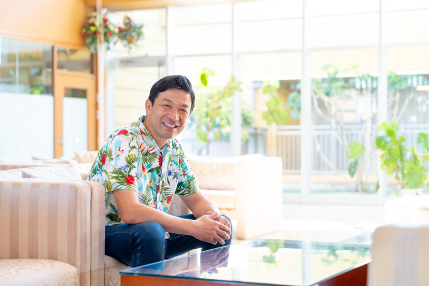 アロハシャツを着て笑顔でソファに座る社会福祉法人征峯会の渡辺和成理事長