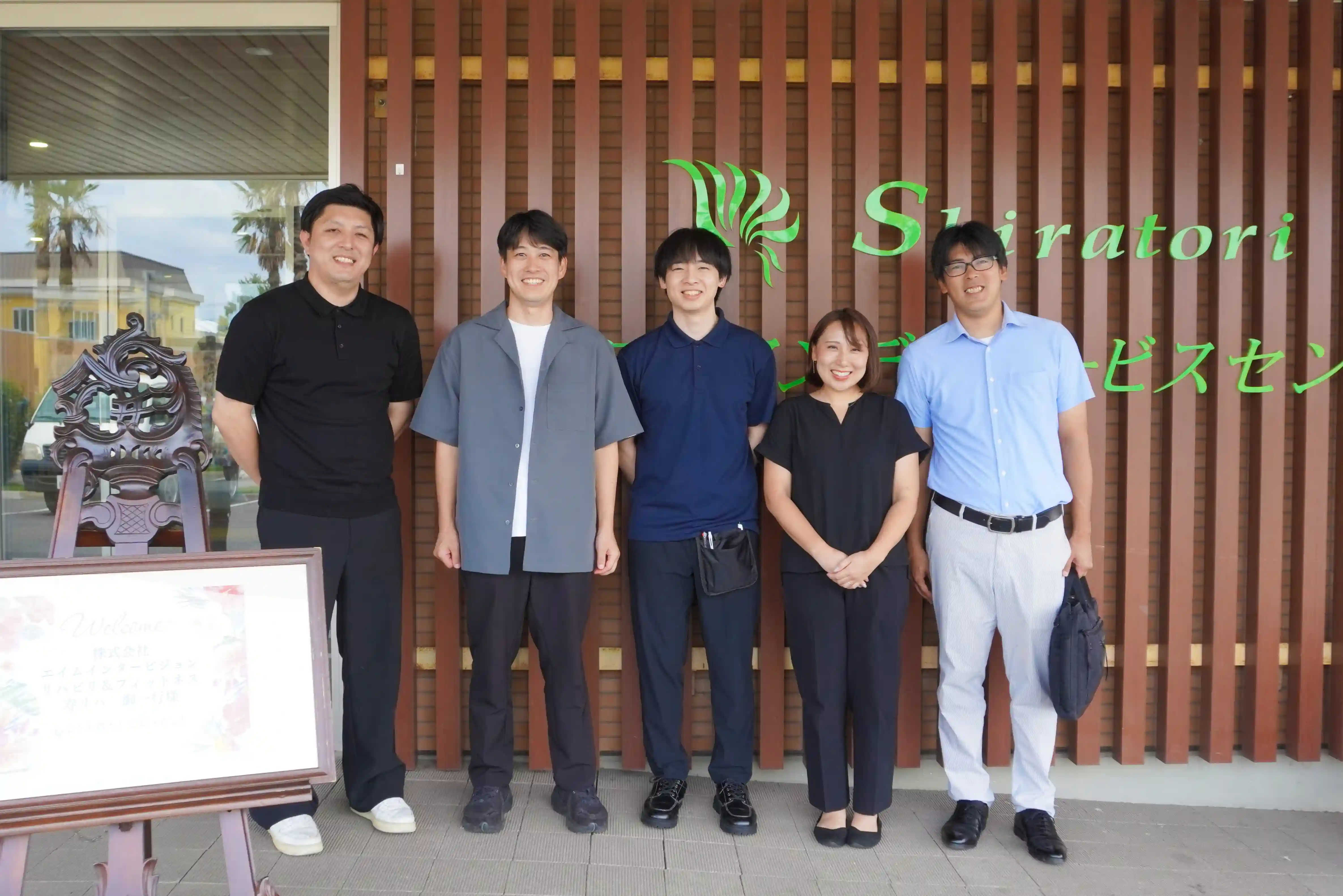 しらとりデイサービスセンターの建物前で並んで笑顔を見せる、株式会社エイムインタービジョンの見学者様と征峯会スタッフの集合写真。