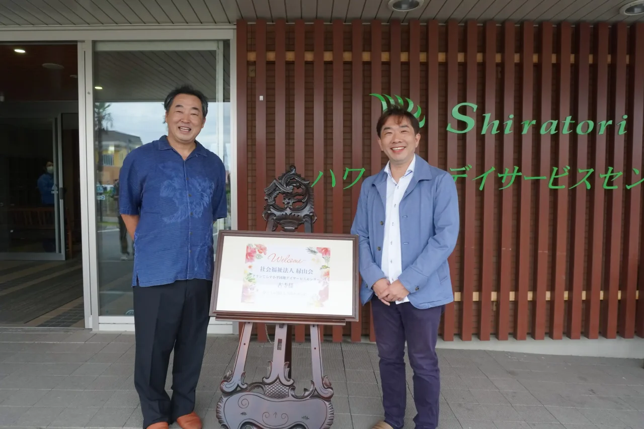 しらとりハワイアンデイデイサービスセンターの看板前でウェルカムボードを囲み笑顔で並ぶ施設長と緑山会の見学者