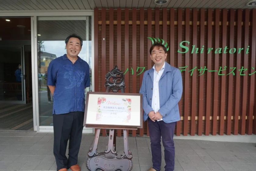しらとりハワイアンデイデイサービスセンターの看板前でウェルカムボードを囲み笑顔で並ぶ施設長と緑山会の見学者